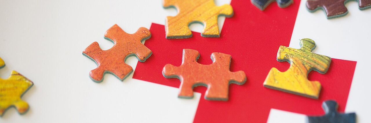 Ein rotes Kreuz ist auf einer weißen Fläche gedruckt. Auf dem Kreuz liegen viele kleine Puzzleteile. Sie sollen das komplexe Zusammenwirken des DRK symbolisieren.