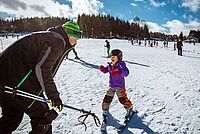 Ein kleines Mädchen fährt auf Skiern. Sie kommt mit großem Lachen auf den Skilehrer zugefahren.
