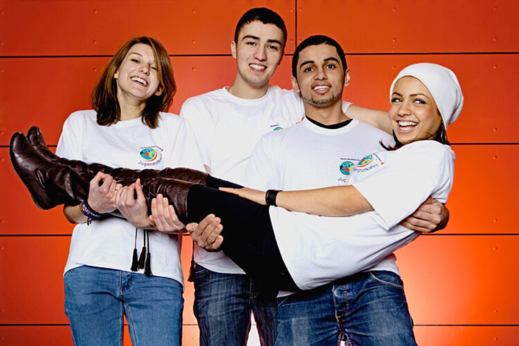 Drei Jugendliche des Jugendrotkreuzes stehen nebeneinander und halten eine Jugendliche quer liegend auf ihren Armen. 