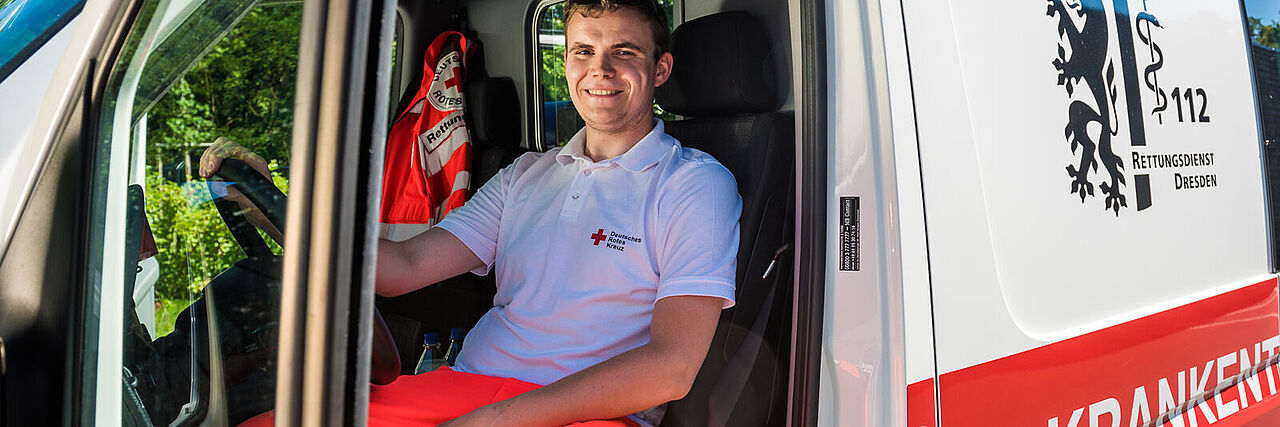 Ein junger Mann sitzt bei offener Tür auf dem Fahrersitz eines Wagens des Krankentransports. Er lächelt in die Kamera und ist bereit für seinen nächsten Einsatz.