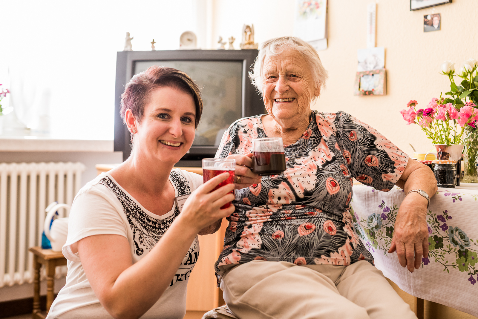 Eine Freiwilligendienstleistende hockt neben einer sitzenden Seniorin. Beide haben ein Saftglas mit einem roten Getränk in der Hand, stoßen an und lächeln in die Kamera.