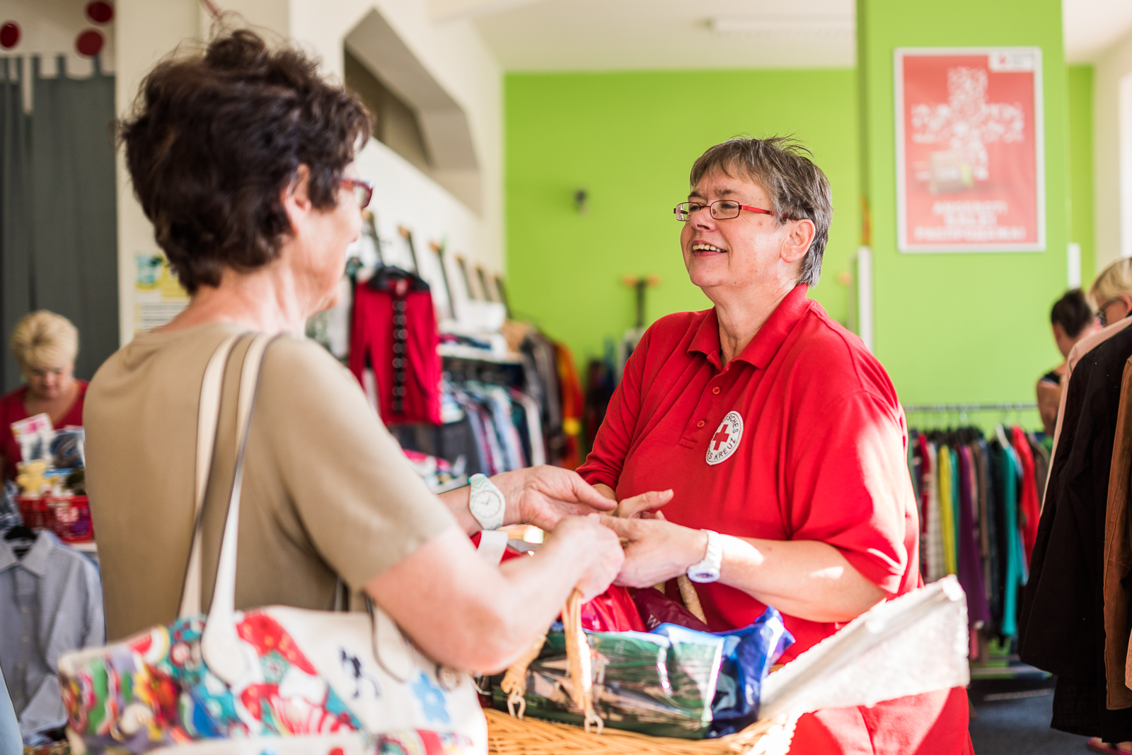 Eine ältere Frau leistet ihren Freiwilligendienst beim Kleiderladen. Sie reicht einer Kundin gerade einen Korb mit ihren gekauften Waren über den Verkaufstresen.