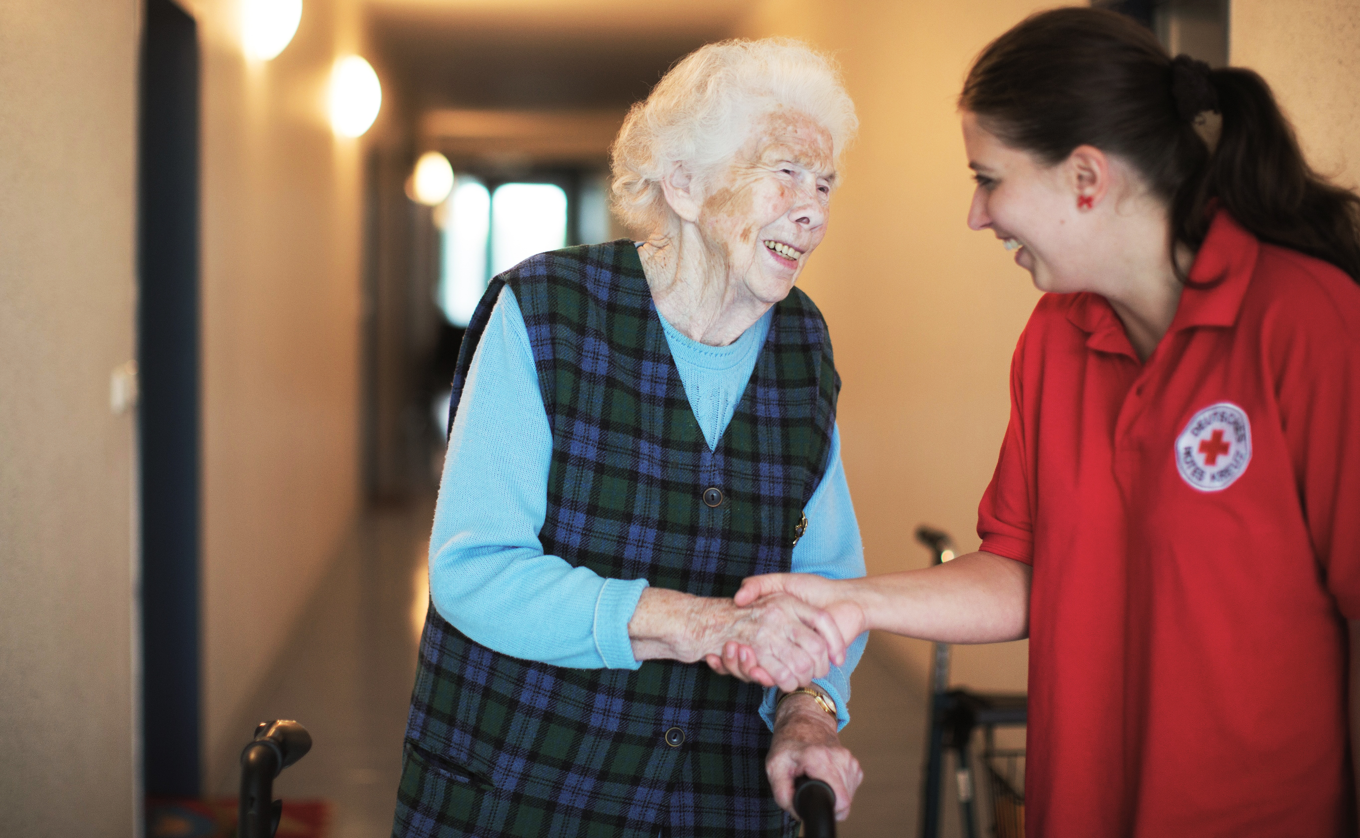 Eine Seniorin greift die Hand ihrer Pflegerin. Die Geste strahlt Vertrauen aus, denn beide lachen miteinander.