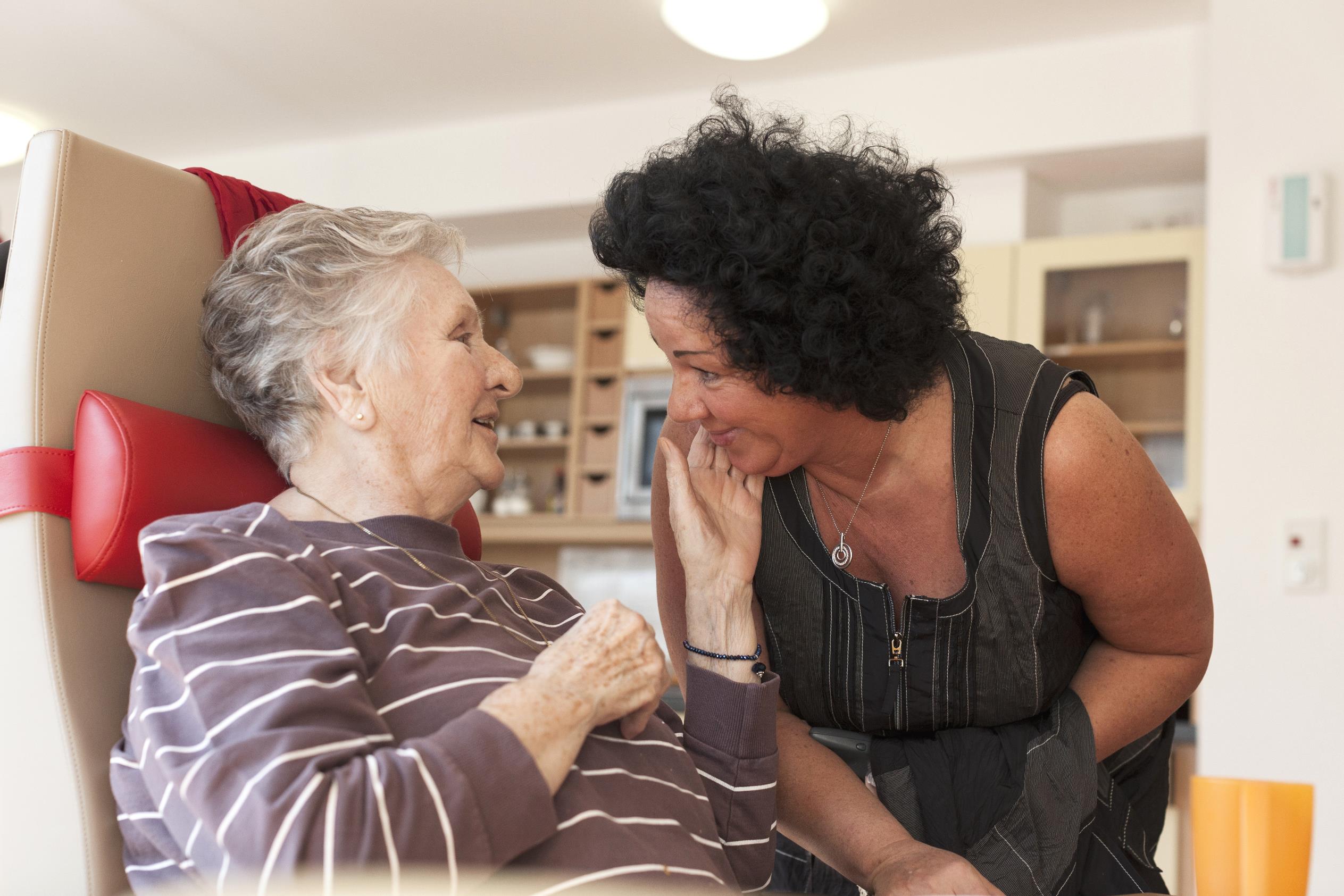 Eine Betreuerin beugt sich zu einer älteren, sitzenden Demenzpatientin herunter. Die ältere Frau streichelt der Betreuerin über die Wange und lächelt.