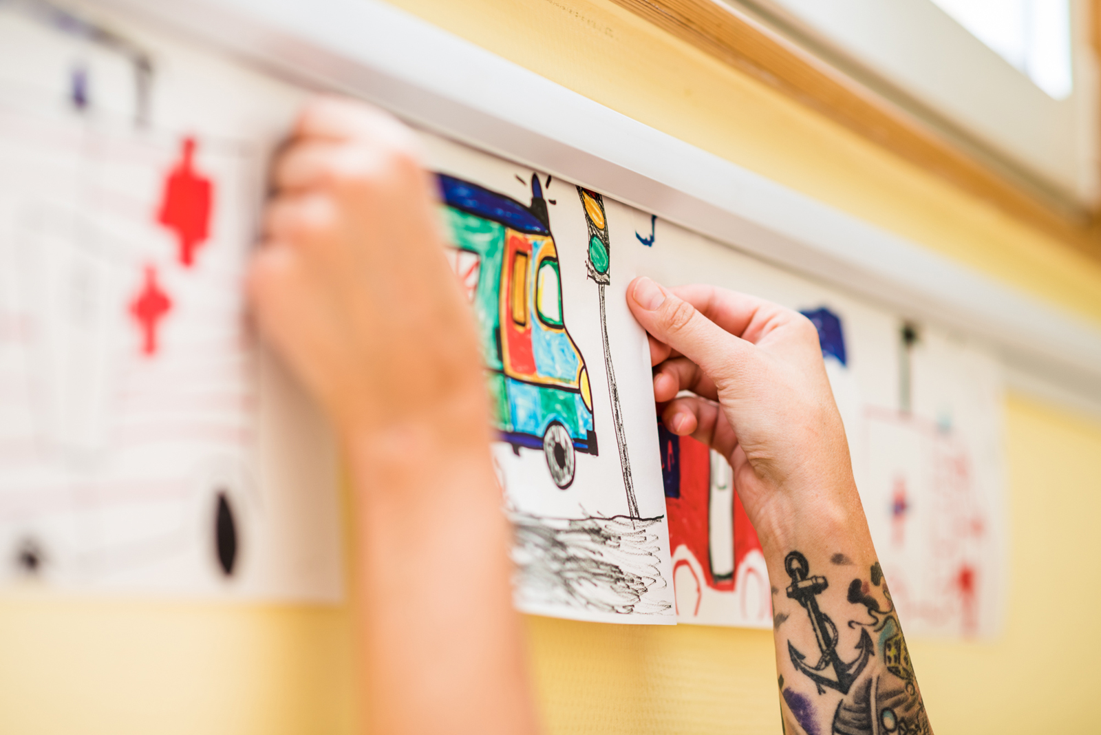 Die Hände von einer erwachsenen Person sind zu sehen. Sie klebt eine Kinderzeichnung an die Wand einer Kita.