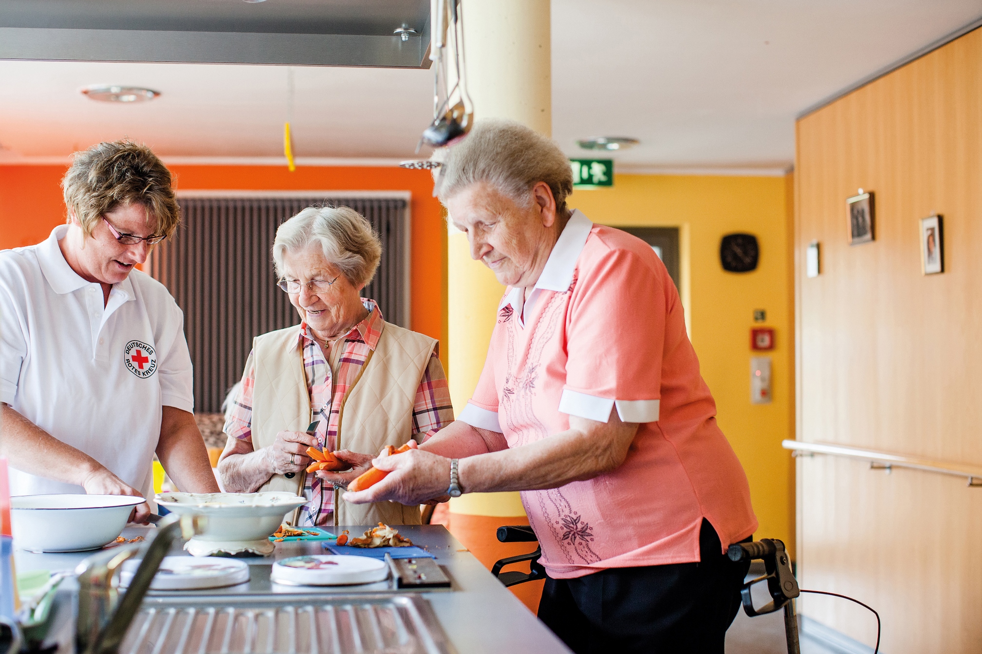Eine DRK Betreuerin und zwei Seniorinnen stehen gemeinsam an einer Küchentheke und schneiden Gemüse klein.