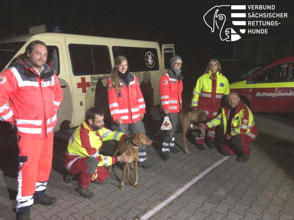 Die Mehrere Personen und ihre Suchhunde der Rettungshundestaffeln Sachsens sind im Einsatz, um vermisste Personen zu finden. Hier stehen sie vor ihrem Einsatzwagen, es ist bereits dunkel.