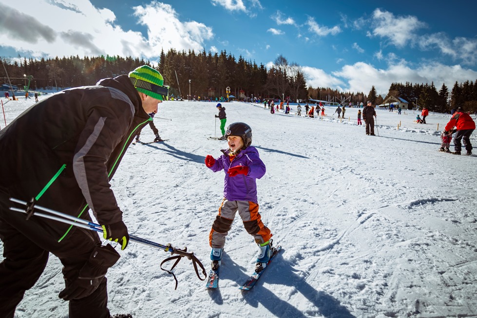 Ein kleines Mädchen fährt auf Skiern. Sie kommt mit großem Lachen auf den Skilehrer zugefahren.