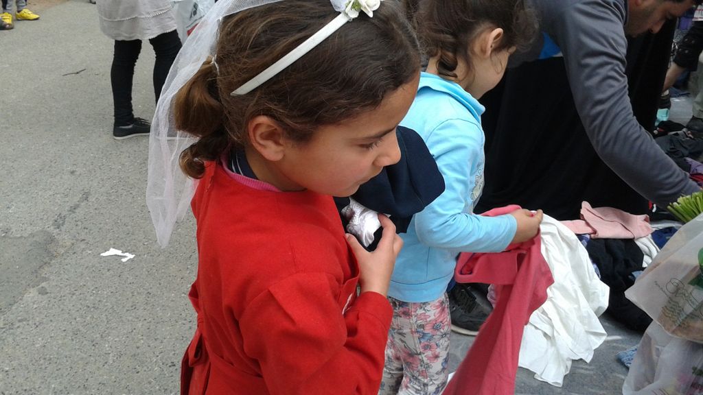 Ein kleines Mädchen aus Syrien steht auf der Straße vor einem Tisch auf dem Kleidung liegt. Das Mädchen trägt einen roten Pullover. 