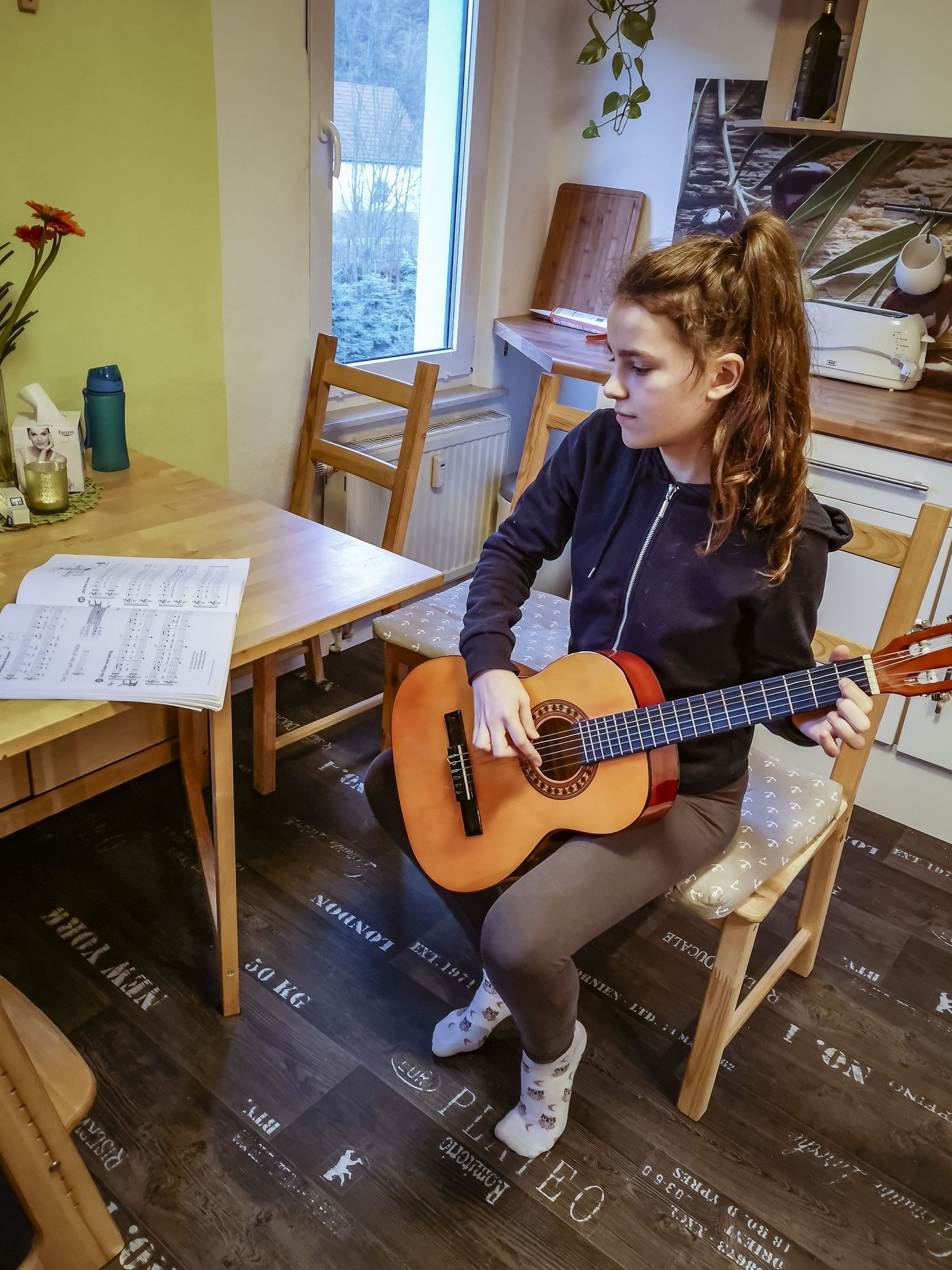 Ein Mädchen sitzt auf einem Stuhl und spielt auf der Gitarre ein Lied. Dabei schaut sie auf ein Notenheft auf dem Tisch.
