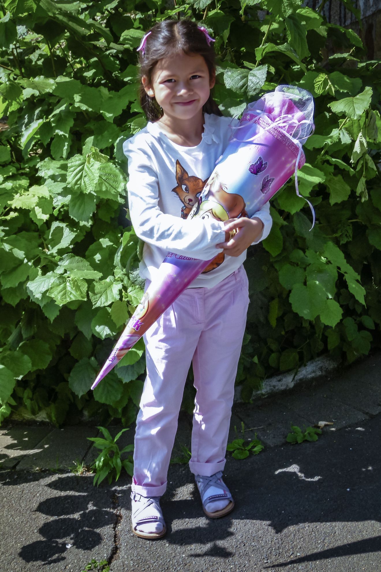 Das dunkelhaarige Mädchen steht vor einer grünen Hecke und hält eine lilafarbene Zuckertüte in der Hand