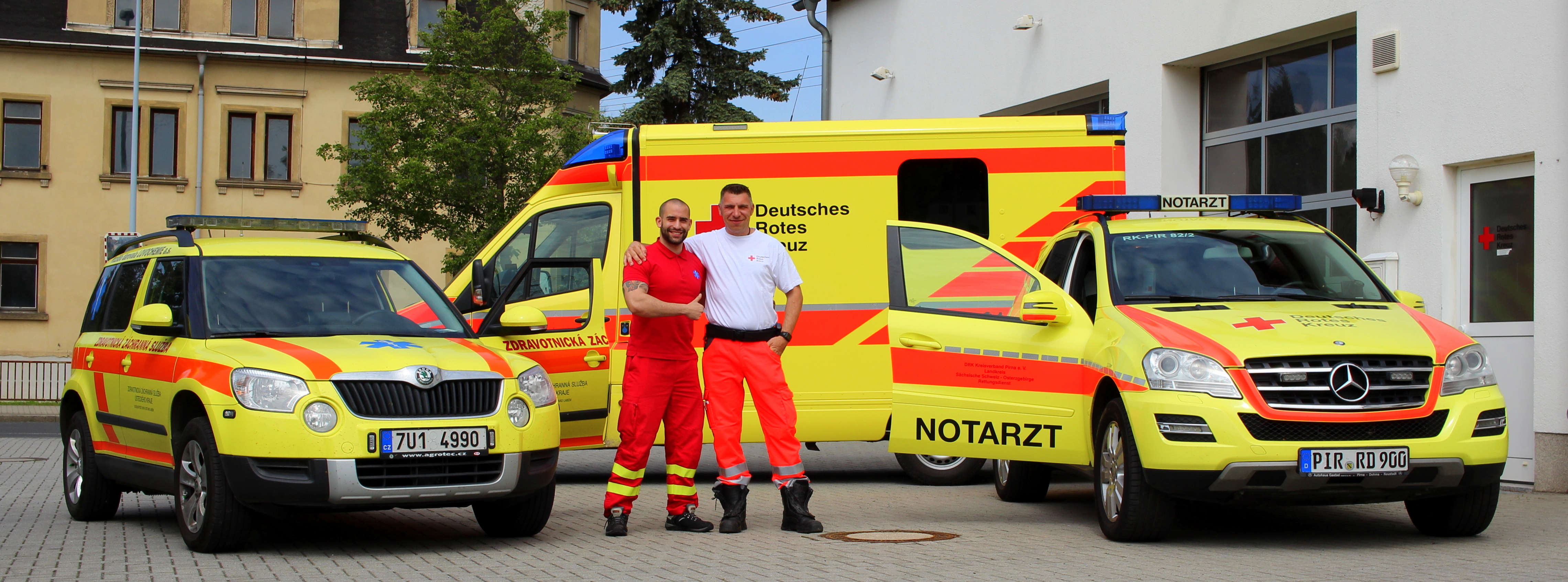 Ein Tschechischer und ein Deutscher Mitarbeiter des Rettungsdienstes stehen Arm in Arm vor ihren Einsatzwagen.