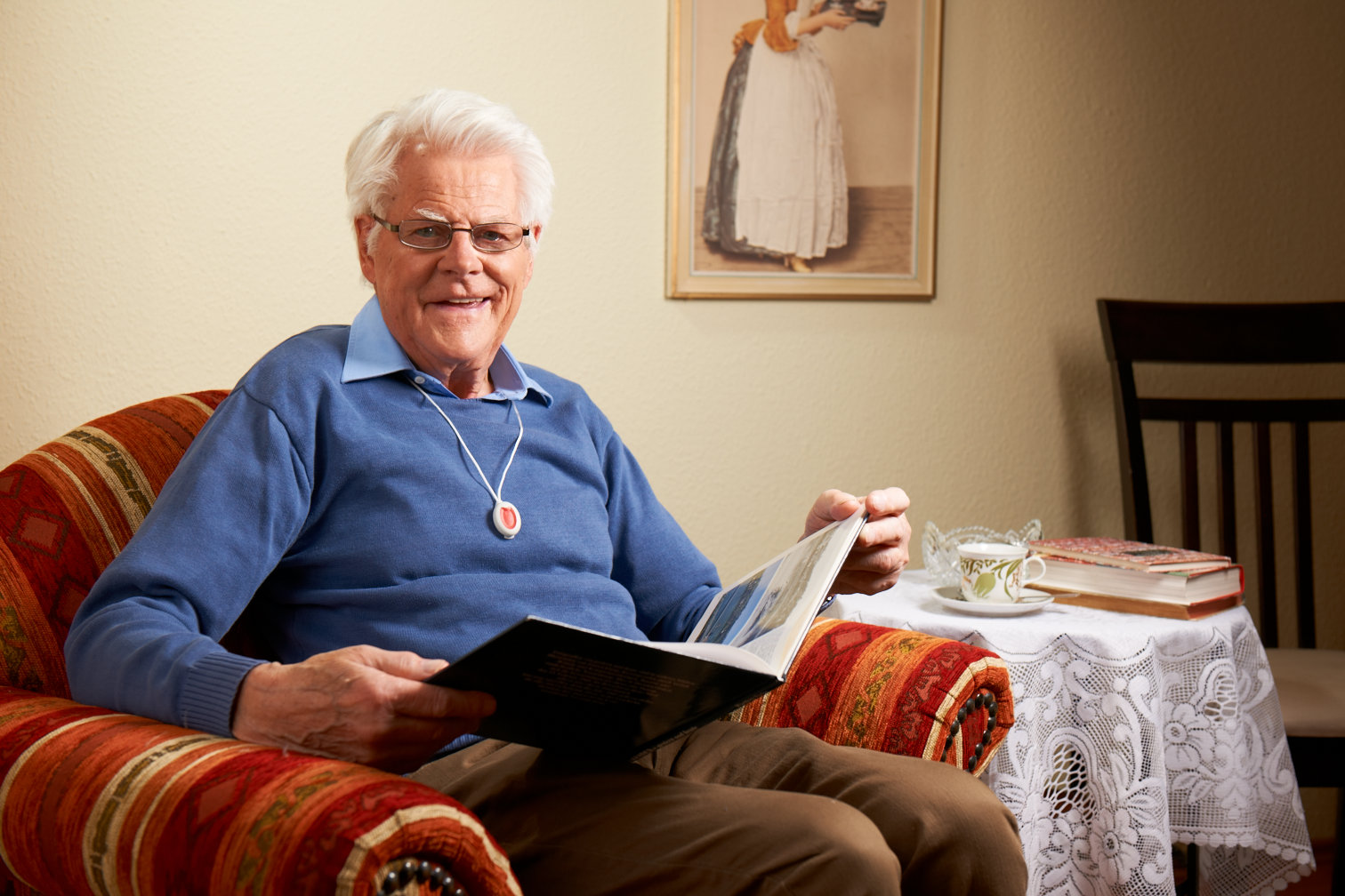 Ein älterer trägt den Notfallknopf des Hausnotrufes an einer Kette um den Hals. Er sitzt in einem Sessel und liest Zeitung.