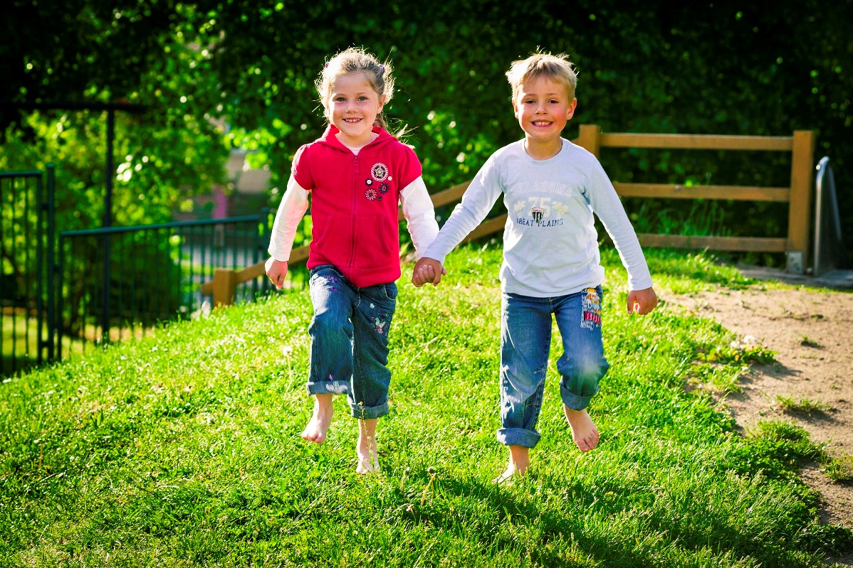 Zwei fröhliche Kinder, ein Junge und ein Mädchen, laufen Hand in Hand über eine Wiese. Sie kommen auf den Kameramann zu und lachen.