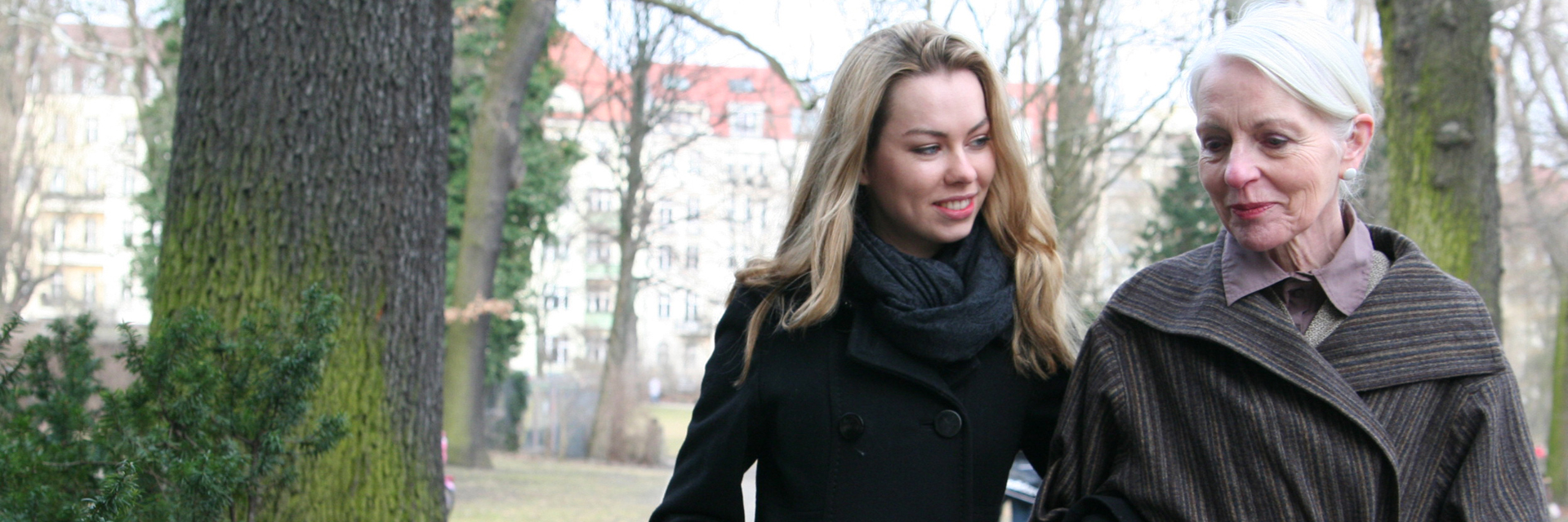 Eine ältere Frau hat ihren Arm bei einer jungen Frau eingehakt. Gemeinsam laufen sie durch einen Park. Im Hintergrund sind Wiesen, Bäume und schließlich ein großes Gebäude zu sehen.