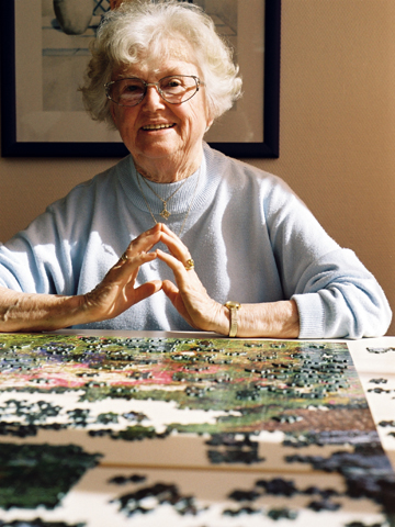Eine Seniorin sitzt an einem Tisch, auf dem ein großes, zum Teil fertiges Puzzle ausgebreitet ist. Sie lächelt in die Kamera.