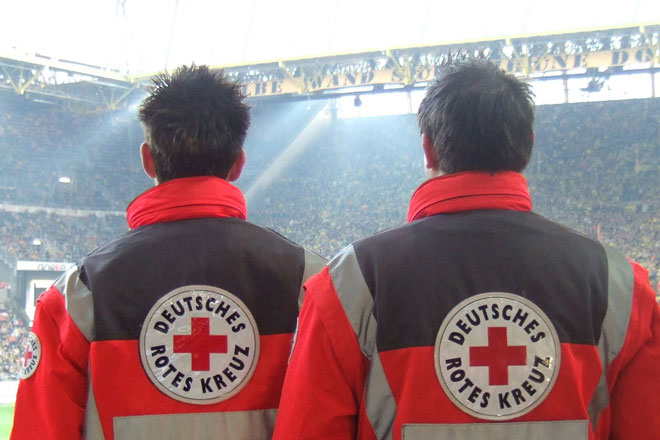 Zwei Rettungssanitäter stehen mit dem Blick in ein mit Menschen gefülltes Stadion mit dem Rücken zur Kamera. Sie sichern eine Großveranstaltung ab.