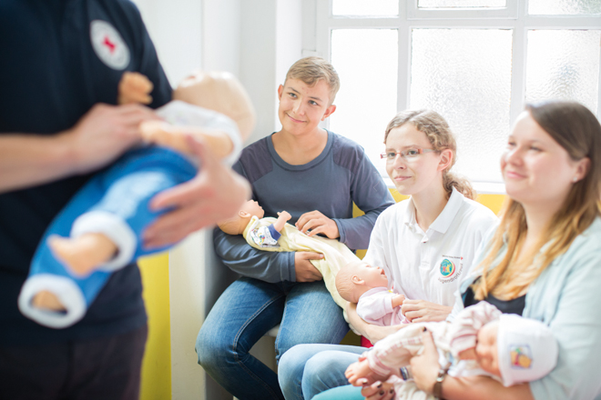 Ein DRK Ausbilder hält eine Babypuppe im Arm und demonstriert den im Hintergund sitzenden Eltern mit ihren Babypuppen auf den Armen, was sie im Ernstfall tun müssen.