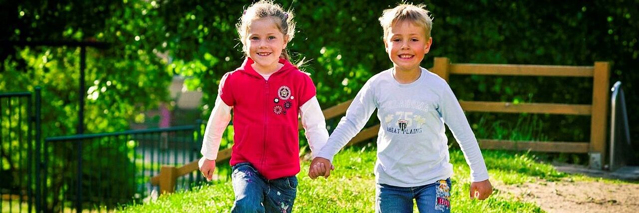 Zwei fröhliche Kinder, ein Junge und ein Mädchen, laufen Hand in Hand über eine Wiese. Sie kommen auf den Kameramann zu und lachen.