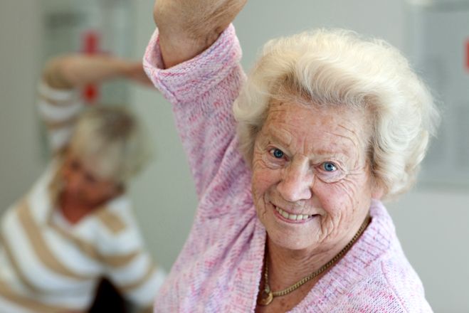 Eine Seniorin streckt ihren rechten Arm über ihren Kopf und lehnt sich nach links, um sich zu strecken. Sie lächelt in die Kamera. Im Hintergrund führt eine weitere Seniorin dieselbe Übung aus.