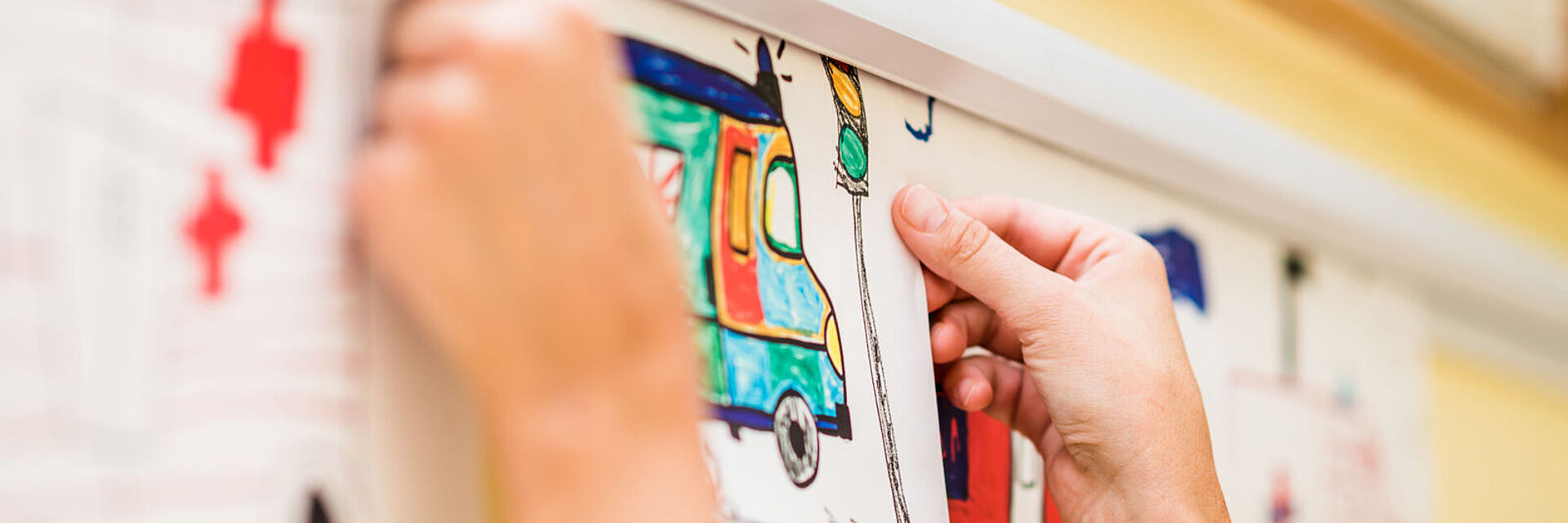 Die Hände von einer erwachsenen Person sind zu sehen. Sie klebt eine Kinderzeichnung an die Wand einer Kita.