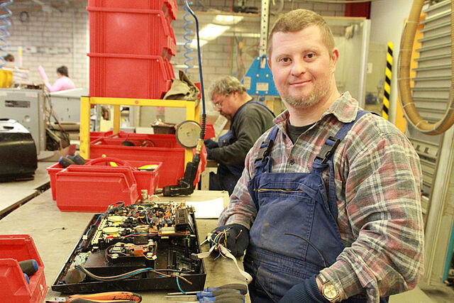 Ein junger Mann steht in einer Werkstatt für Menschen mit Behinderung. Er trägt eine blaue Arbeitshose und arbeitet an einer Werkbank.