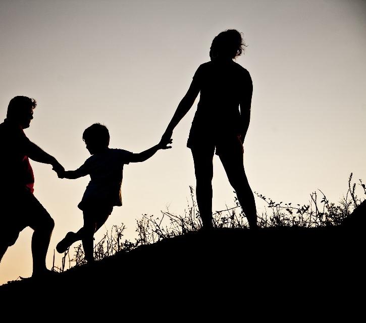 Familienerholung Ein Paar hält ein Kind in seiner Mitte an den Händen. Sie gehen einen Berg hinauf. Es sind nur Silhouetten zu sehen.