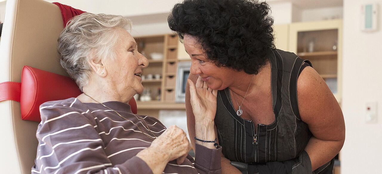 Eine Betreuerin beugt sich zu einer älteren, sitzenden Demenzpatientin herunter. Die ältere Frau streichelt der Betreuerin über die Wange und lächelt.