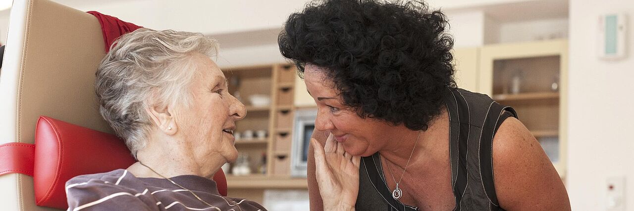 Eine Betreuerin beugt sich zu einer älteren, sitzenden Demenzpatientin herunter. Die ältere Frau streichelt der Betreuerin über die Wange und lächelt.