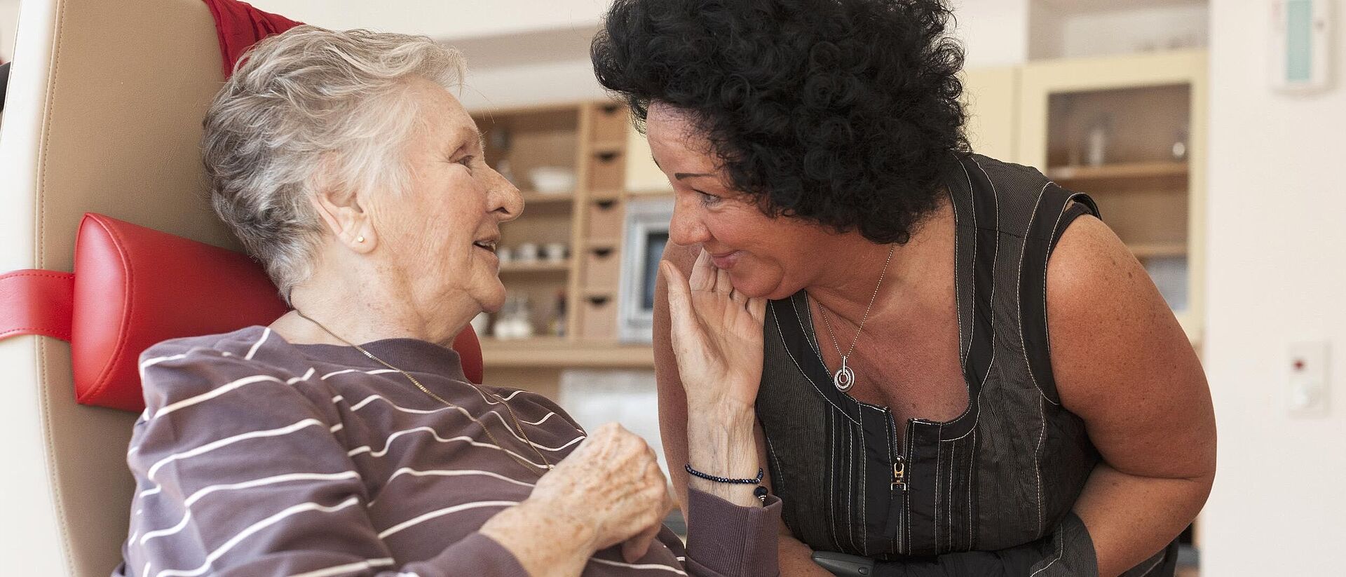 Eine Betreuerin beugt sich zu einer älteren, sitzenden Demenzpatientin herunter. Die ältere Frau streichelt der Betreuerin über die Wange und lächelt.