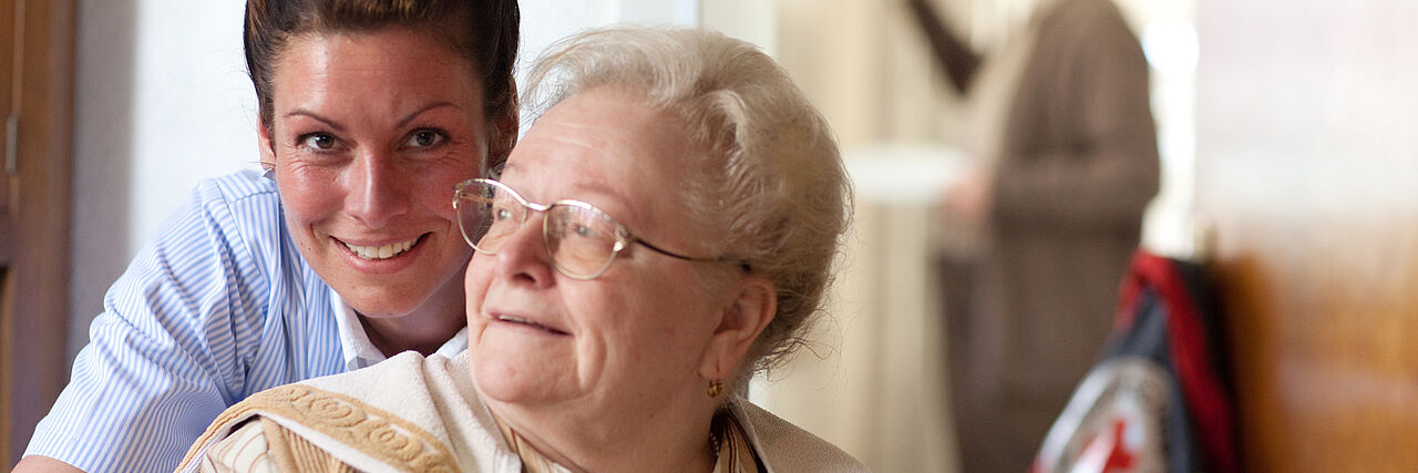Eine Pflegerin steht hinter einer sitzenden Seniorin und umarmt sie. Die Seniorin dreht sich zu ihr und lächelt.