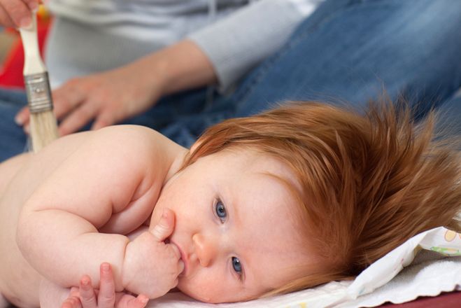 Ein Baby liegt auf dem Bauch, den Kopf zur Seite gedreht. Der Oberkörper ist nackt und eine Person im Hintergrund fährt in entspannenden Bewegungen mit einem sauberen Pinsel über den Rücken des Kindes.