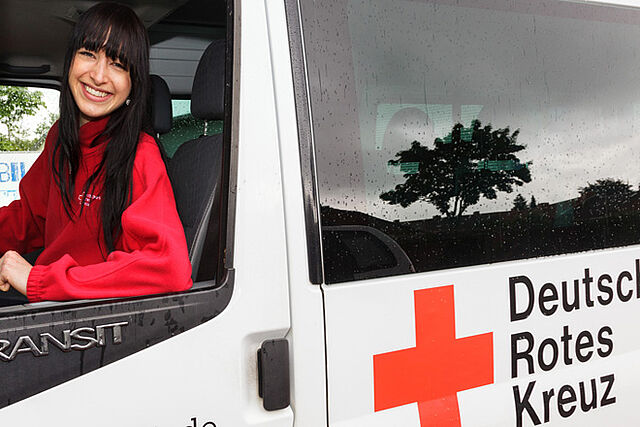 Eine DRK Mitarbeiterin des Fahrdienstes sitzt im Fahrzeug und schaut lächelnd aus dem Fahrerfenster.