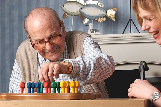 Eine Pflegerin spielt mit einem Senior ein Brettspiel. Er ist gerade am Zug und die beiden lachen über etwas.