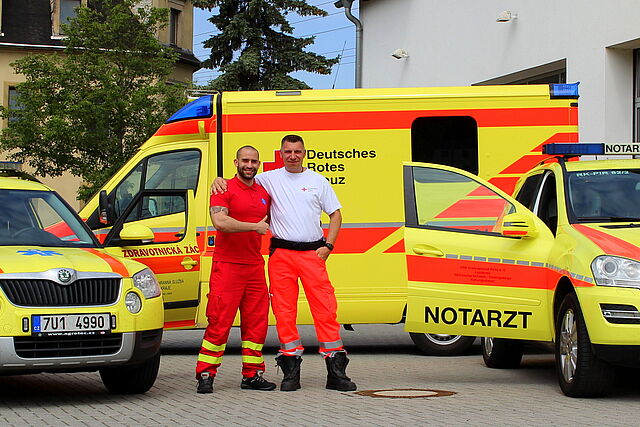 Ein Tschechischer und ein Deutscher Mitarbeiter des Rettungsdienstes stehen Arm in Arm vor ihren Einsatzwagen.