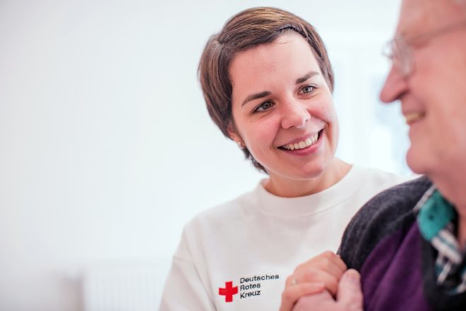 Eine DRK Mitarbeiterin stützt einen Senioren. Beide lächeln sich an.