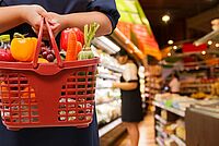 Eine Person im Vordergrund hält einen vollen Einkaufskorb. Im Hintergrund sieht man einen Gang im Supermarkt.