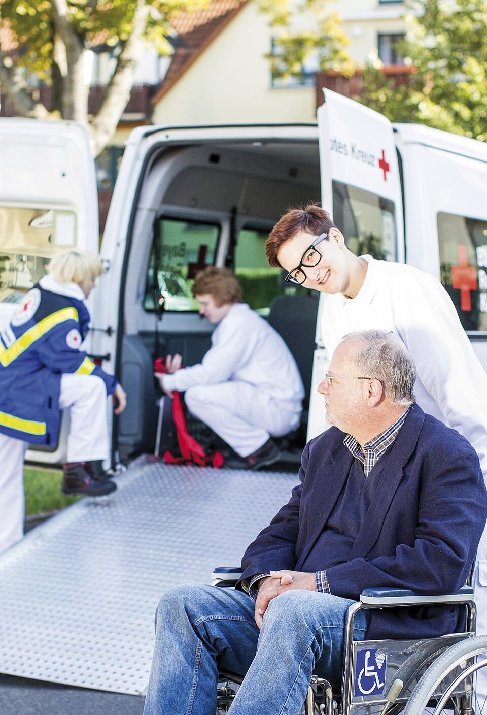 Ein DRK Betreuer schiebt den Rollstuhl eines älteren Mannes. Hinter Ihnen bereiten zwei Mitarbeiterinnen den Transporter für die Fahrt vor. Der Transporter hat eine Rampe für den Rollstuhl.