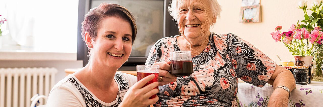 Eine Freiwilligendienstleistende hockt neben einer sitzenden Seniorin. Beide haben ein Saftglas mit einem roten Getränk in der Hand, stoßen an und lächeln in die Kamera.