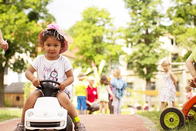 Ein kleines Mädchen fährt mit einem Bobbycar über das Gelände des Kindergartens. Im Hintergrund sieht man Kinder spielen und mit einer Erzieherin sprechen.