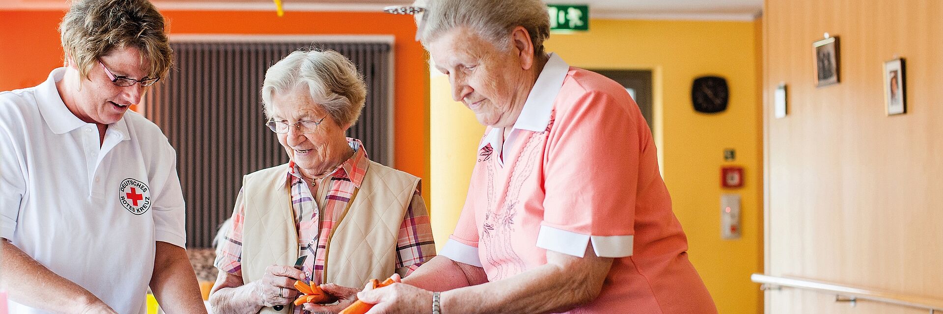 Eine DRK Betreuerin und zwei Seniorinnen stehen gemeinsam an einer Küchentheke und schneiden Gemüse klein.