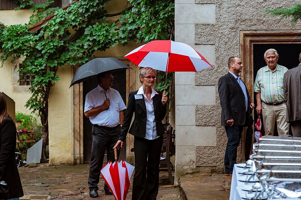 Eine Mitarbeiterin des DRK hält Schirme für die ankommenden Gäste bereit. Sie hält selbst einen Schirm und stützt sich auf einen zweiten.