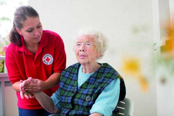 Eine junge DRK Pflegerin hält die Hand einer auf einem Stuhl sitzenden Seniorin.