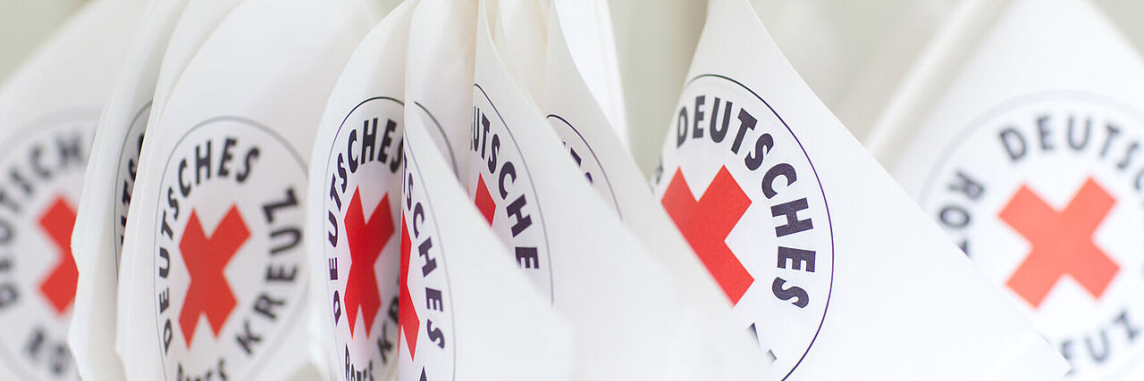 Mehrere Fähnchen mit dem Logo des Deutschen Roten Kreuzes sind nebeneinander zu sehen. 