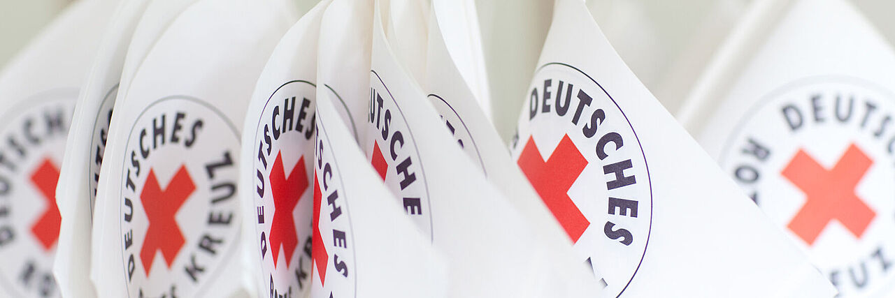 Mehrere Fähnchen mit dem Logo des Deutschen Roten Kreuzes sind nebeneinander zu sehen. 