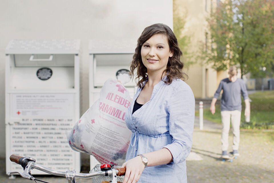 Eine Frau steht am Kleidercontainer und wirft eine Tüte mit aussortierter Kleidung ein.