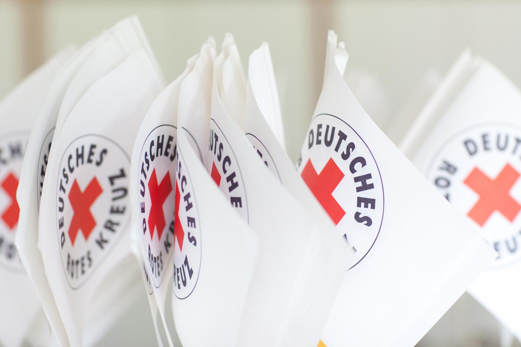 Mehrere Fähnchen mit dem Logo des Deutschen Roten Kreuzes sind nebeneinander zu sehen. 