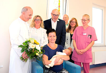 Claire Krumbiegel brachte die 1000. Geburt des DRK Krankenhauses hinter sich und einen kleinen Jungen auf die Welt. Auf dem Bild posieren Ärzte, Schwestern, die Mutter mit ihrem Baby sowie das DRK Vorstandsmitglied Kerstin Trautmann. Insgesamt sechs Personen stehen um die Mutter, die mit ihrem Sohn in der Mitte sitzt.