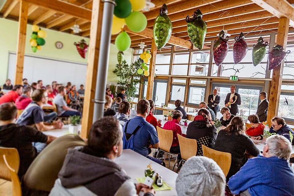 Die Mitarbeiter der Werkstätten Meißen sitzen in einem großen Saal und schauen zum ehemaligen und zum neuen Werkstattleiter.