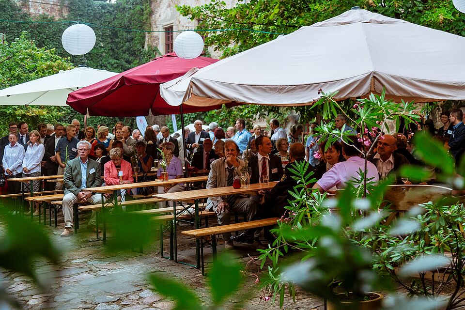 Im Schlosshof stehen Bierbänke und Biertische, große Schirme bieten Schutz vor Sonne und Regen, Lampions beleuchten den Hof. Viele Menschen sitzen gemütlich beieinander.